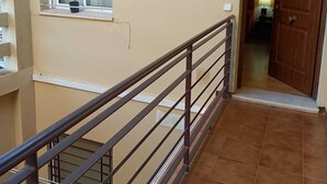 Un couloir doté d’un sol carrelé, une fenêtre avec un rideau, une porte marquée de la lettre « D » et une rambarde en métal.