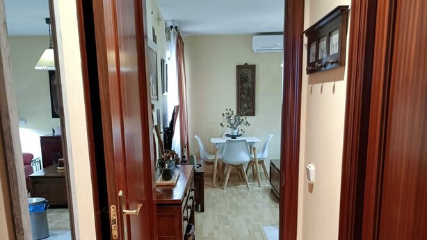 Un couloir avec des portes en bois, un coin repas avec une table et des chaises blanches, et une cuisine équipée de placards et d’une cuisinière.