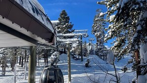 Une station de télésiège enneigée, avec un téléphérique, entourée d’arbres chargés de neige et sous un ciel bleu dégagé.