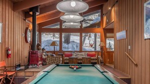 Une table de billard dans une pièce en bois, avec vue sur un paysage enneigé par de grandes fenêtres.