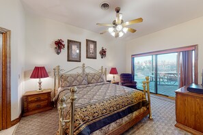 A bedroom with a large bed, two bedside tables with lamps, a ceiling fan, and a view of the outdoors through a large window.
