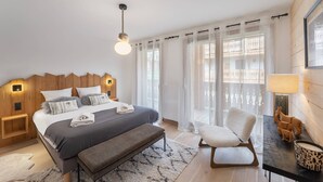 Et moderne soveværelse med sengegavl i træ, en stor seng med gråt og hvidt dynebetræk, en bænk, en stol og et lille bord med en lampe.