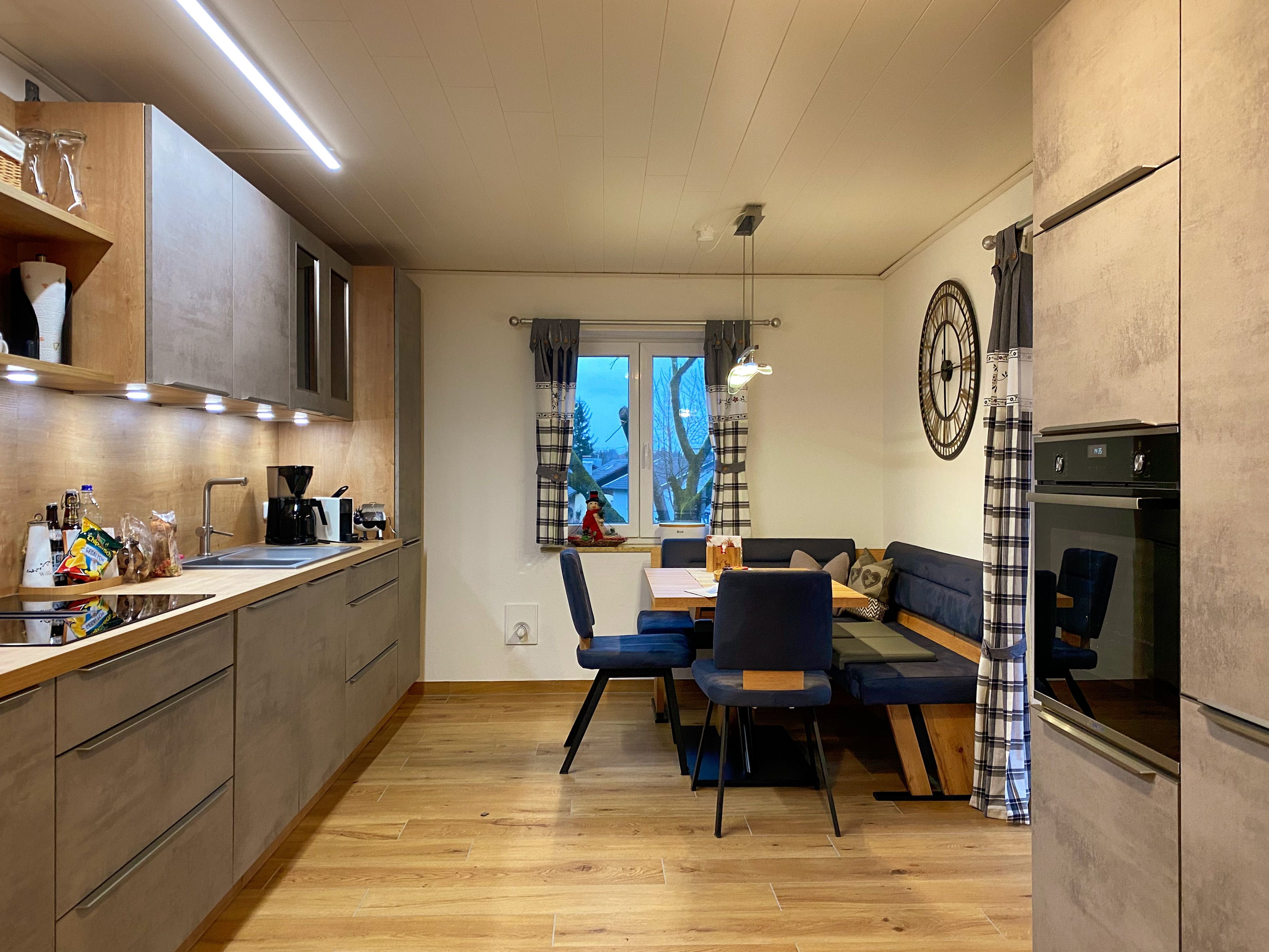 Eine moderne Küche mit Holzschränken, ein Essbereich mit blauem Sofa und Stühlen, sowie ein großes Fenster mit Vorhängen.