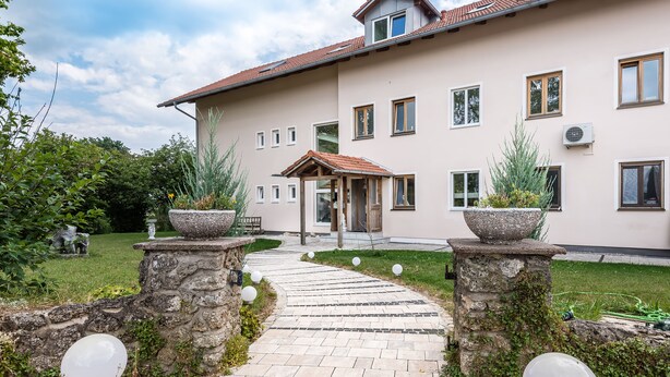 Un bâtiment avec un chemin pavé menant à une entrée en bois, des piliers en pierre ornés de motifs sphériques et un jardin bien entretenu.