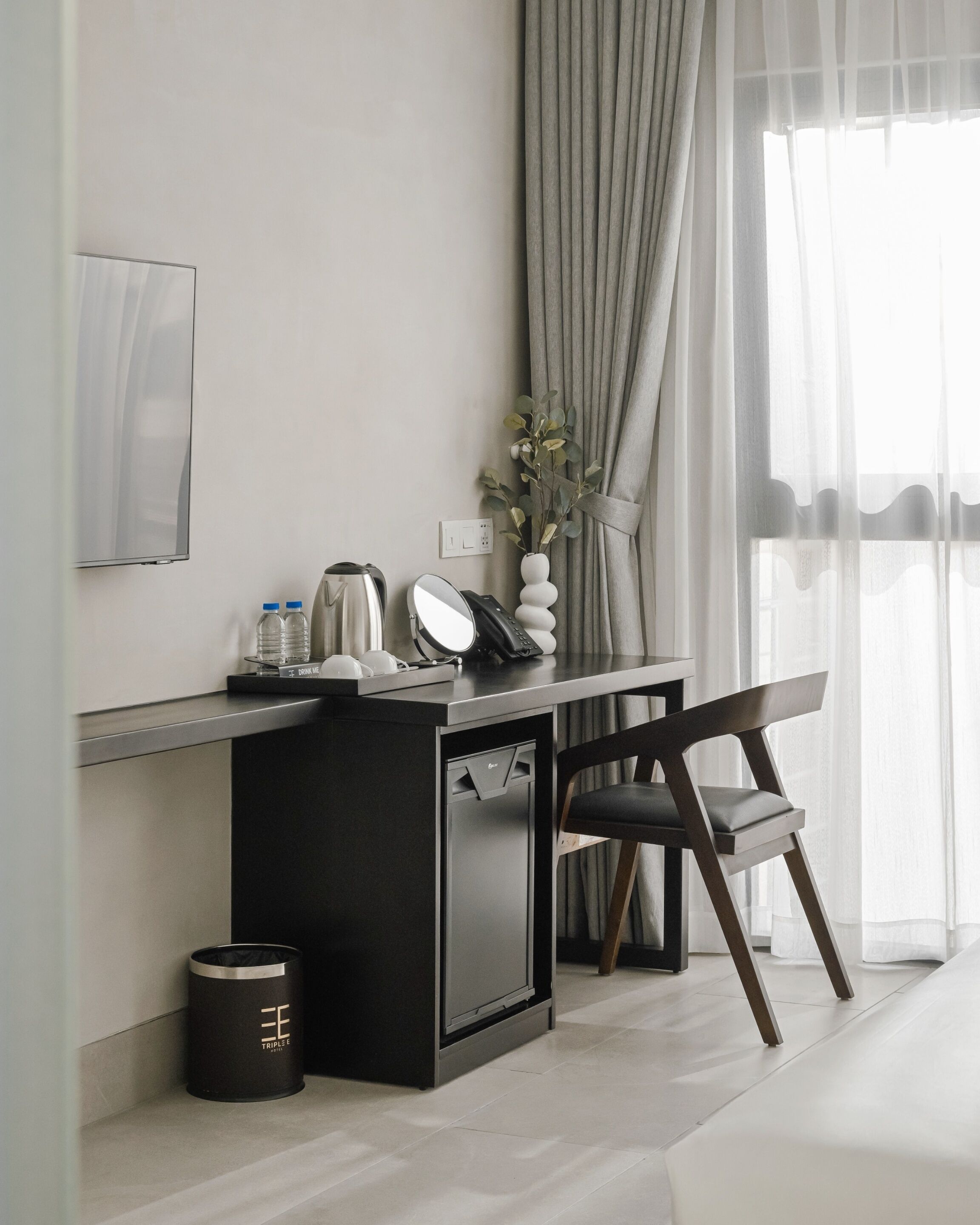 Une chambre d’hôtel moderne avec un bureau minimaliste, une chaise, une petite poubelle, une bouteille d’eau, une bouilloire et un vase avec des feuilles vertes.