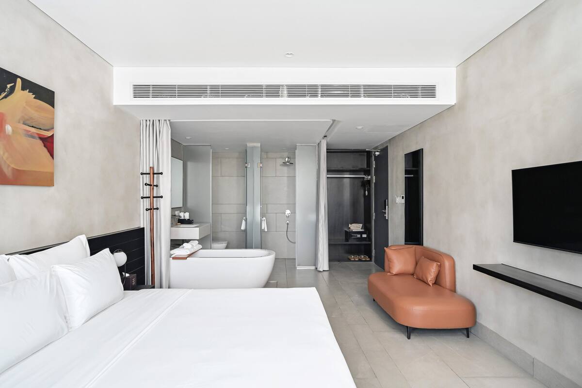 A modern hotel room with a large bed, a bathtub, a shower area, a seating area, and a mounted TV.