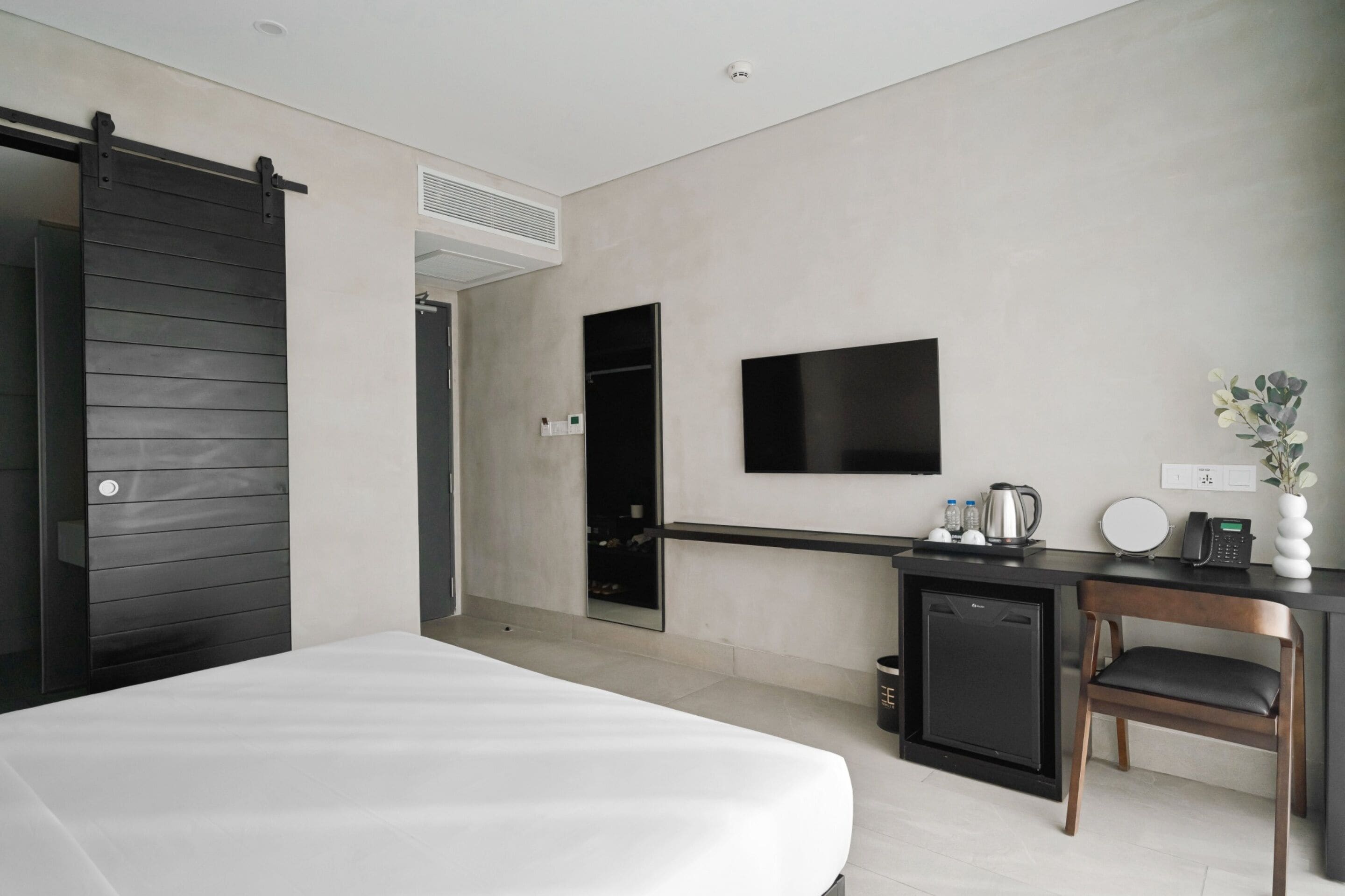 A modern hotel room with a bed, a desk with a telephone and a small cabinet, a television mounted on the wall, and a sliding door.