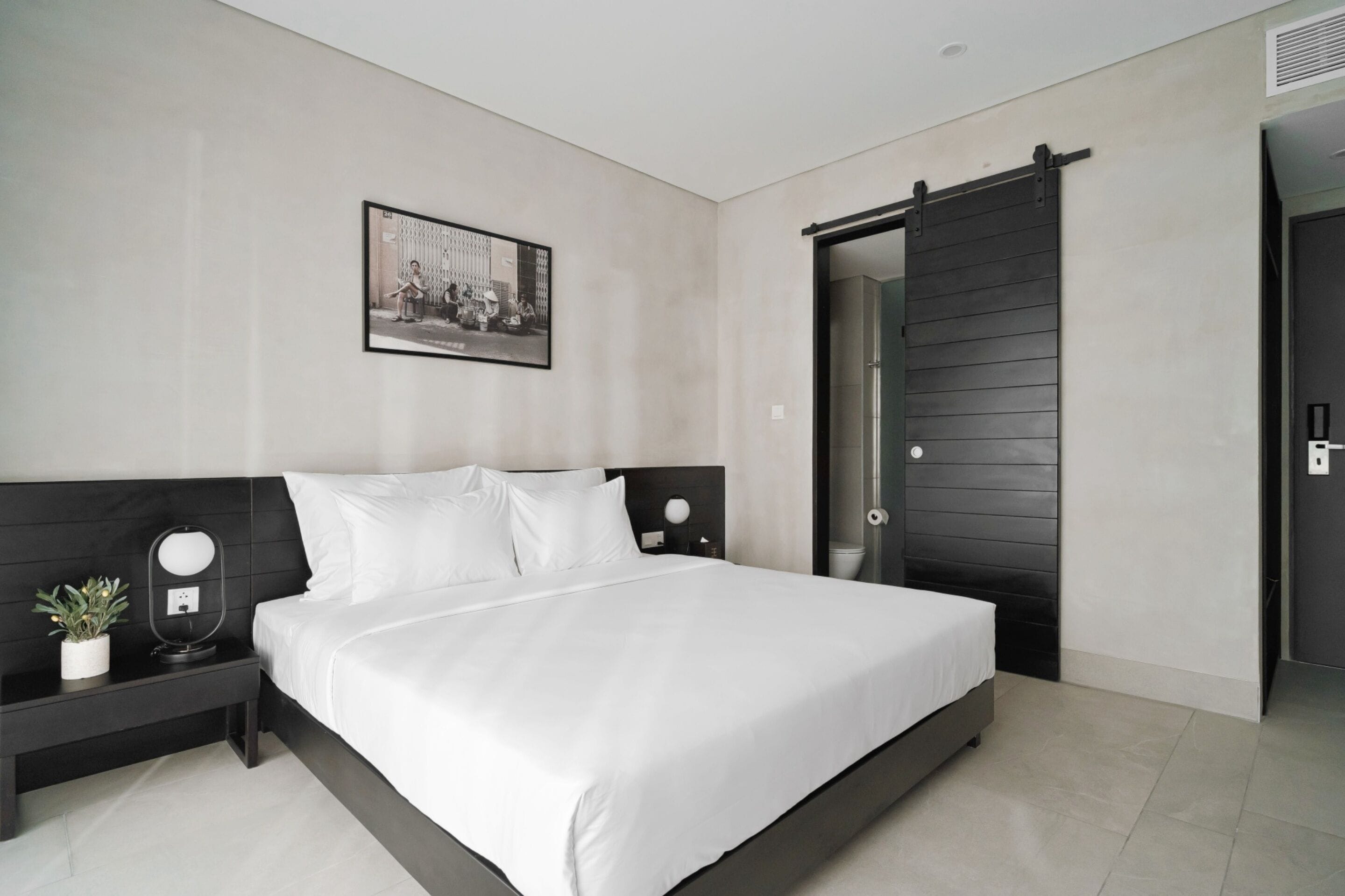 A modern bedroom with a large bed, a nightstand with a mirror, a potted plant, and a sliding door.