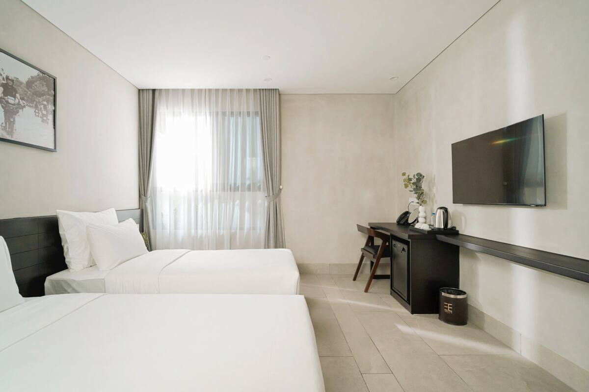 A modern hotel room with two beds, a desk with a chair, a TV mounted on the wall, and a window with sheer curtains.