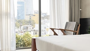 Un dormitorio moderno con una cama grande, una mesita de noche pequeña y una silla. Se aprecia una vista panorámica de la ciudad a través de amplios ventanales.