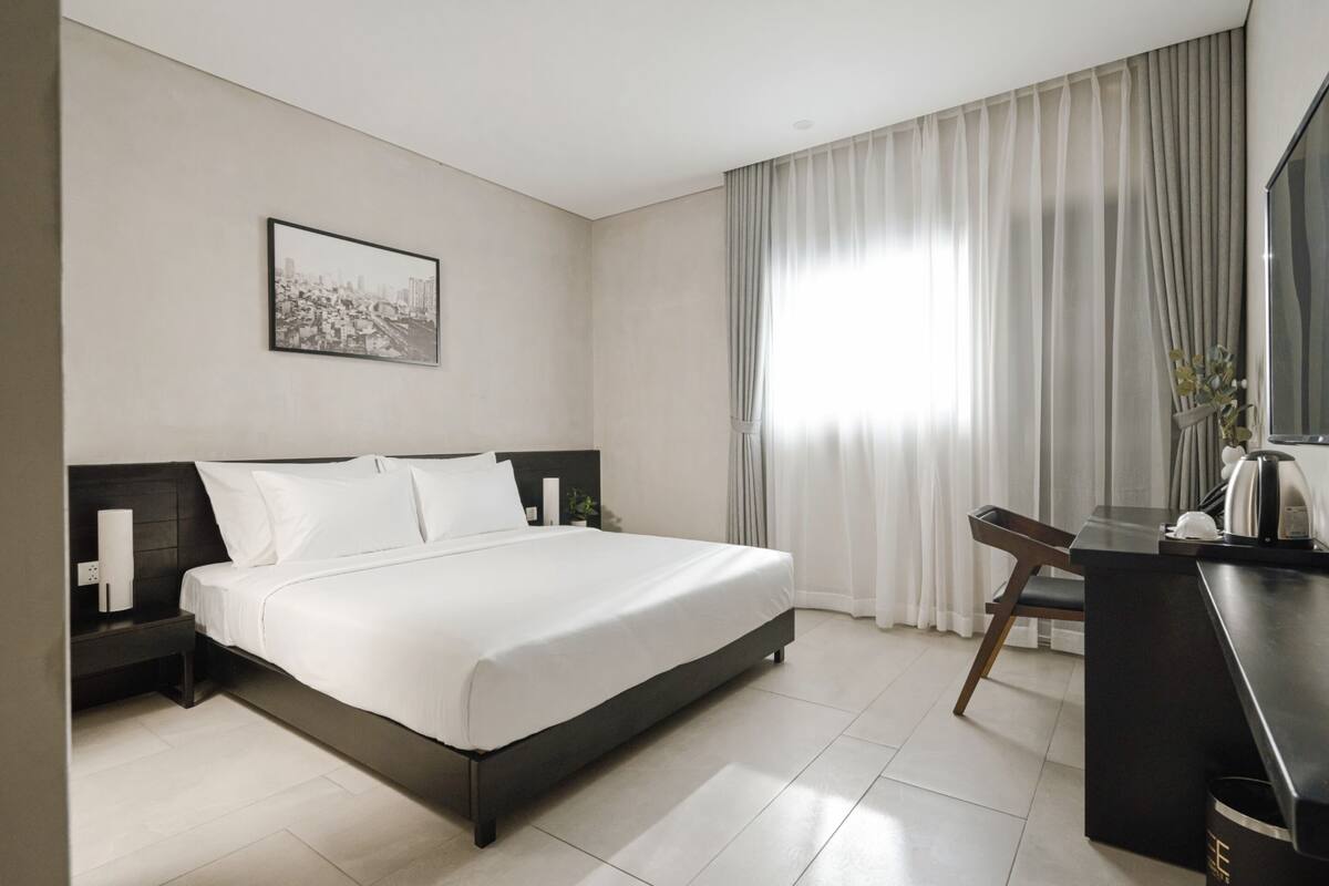 A modern hotel room with a large bed, a desk with a chair, a flat-screen TV, and a cityscape painting on the wall.