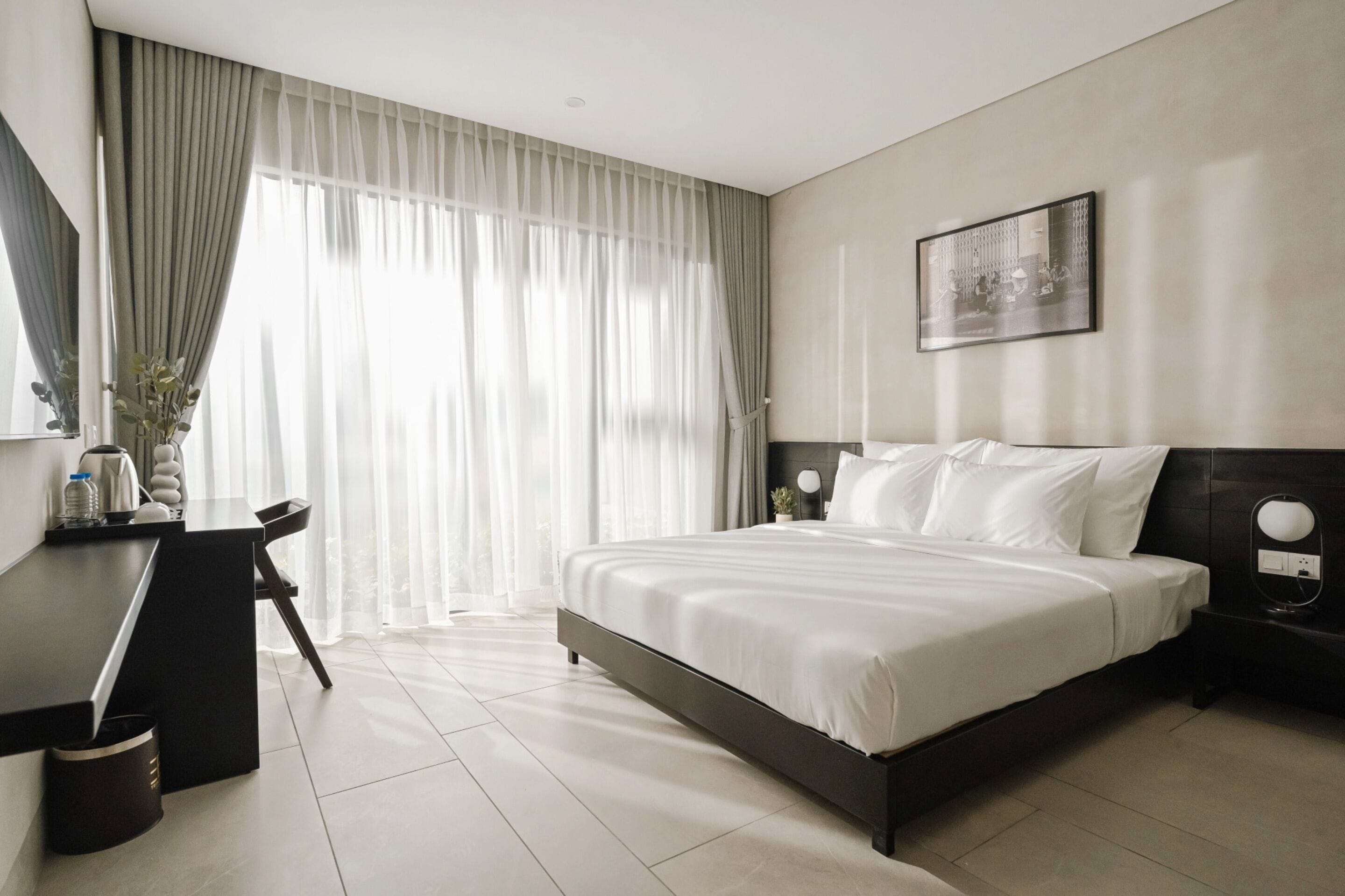 Une chambre d’hôtel moderne avec un grand lit, un bureau et une chaise. Il y a une fenêtre avec des rideaux légers et un tableau encadré au mur.