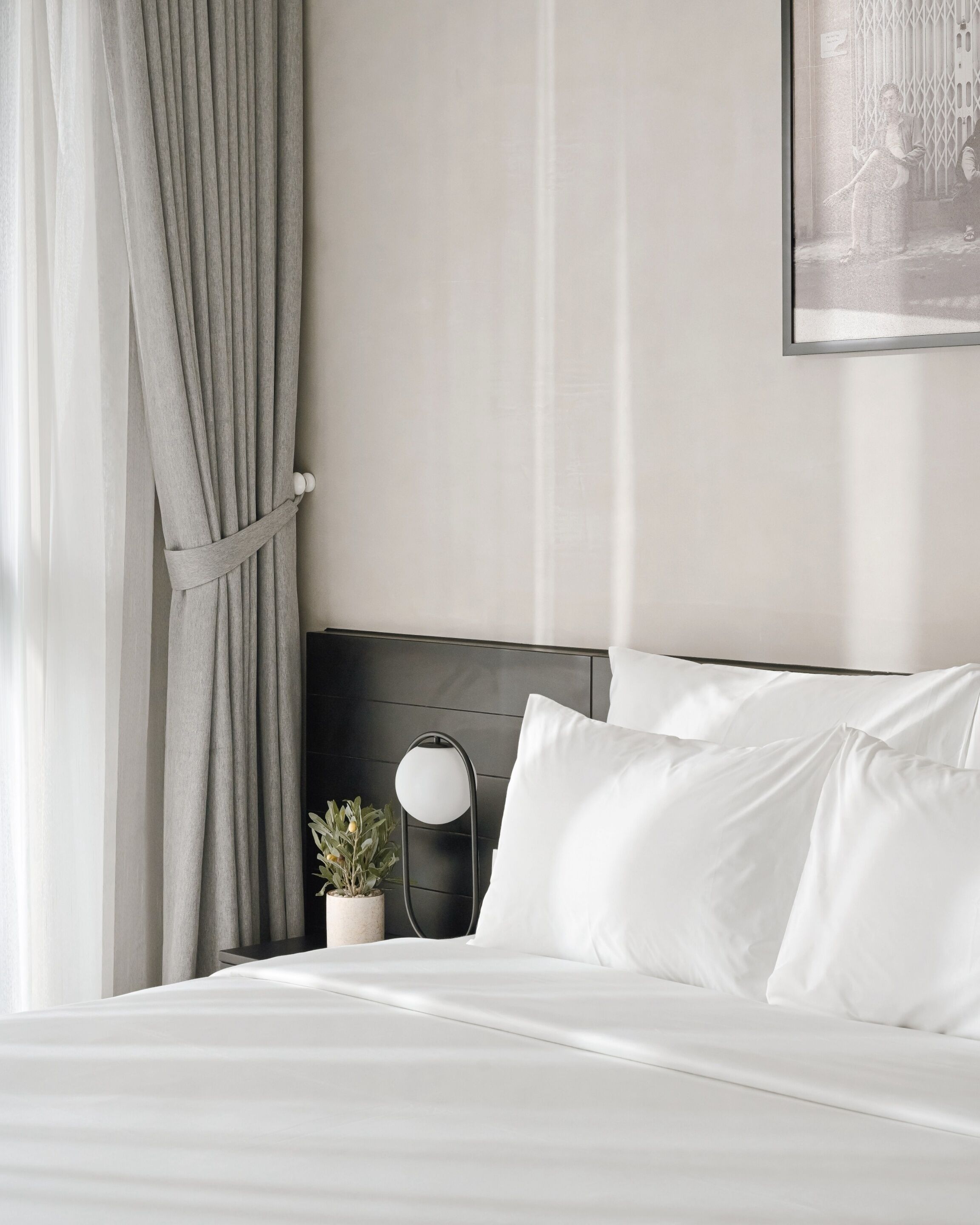 A neatly made bed with white linens, a dark headboard, and a bedside table with a lamp and a small plant.