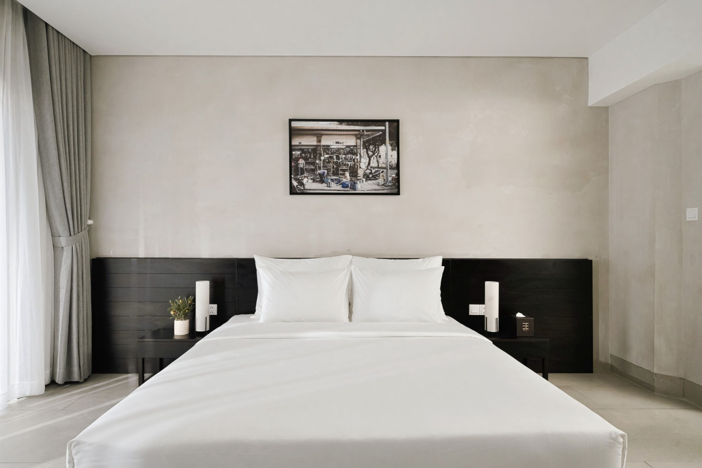 A modern hotel room with a large bed, a headboard with a black panel, a bedside table with a lamp, and a framed picture on the wall.