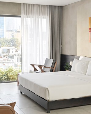 A modern bedroom with a large bed, a chair, and a view of the city through sheer curtains.