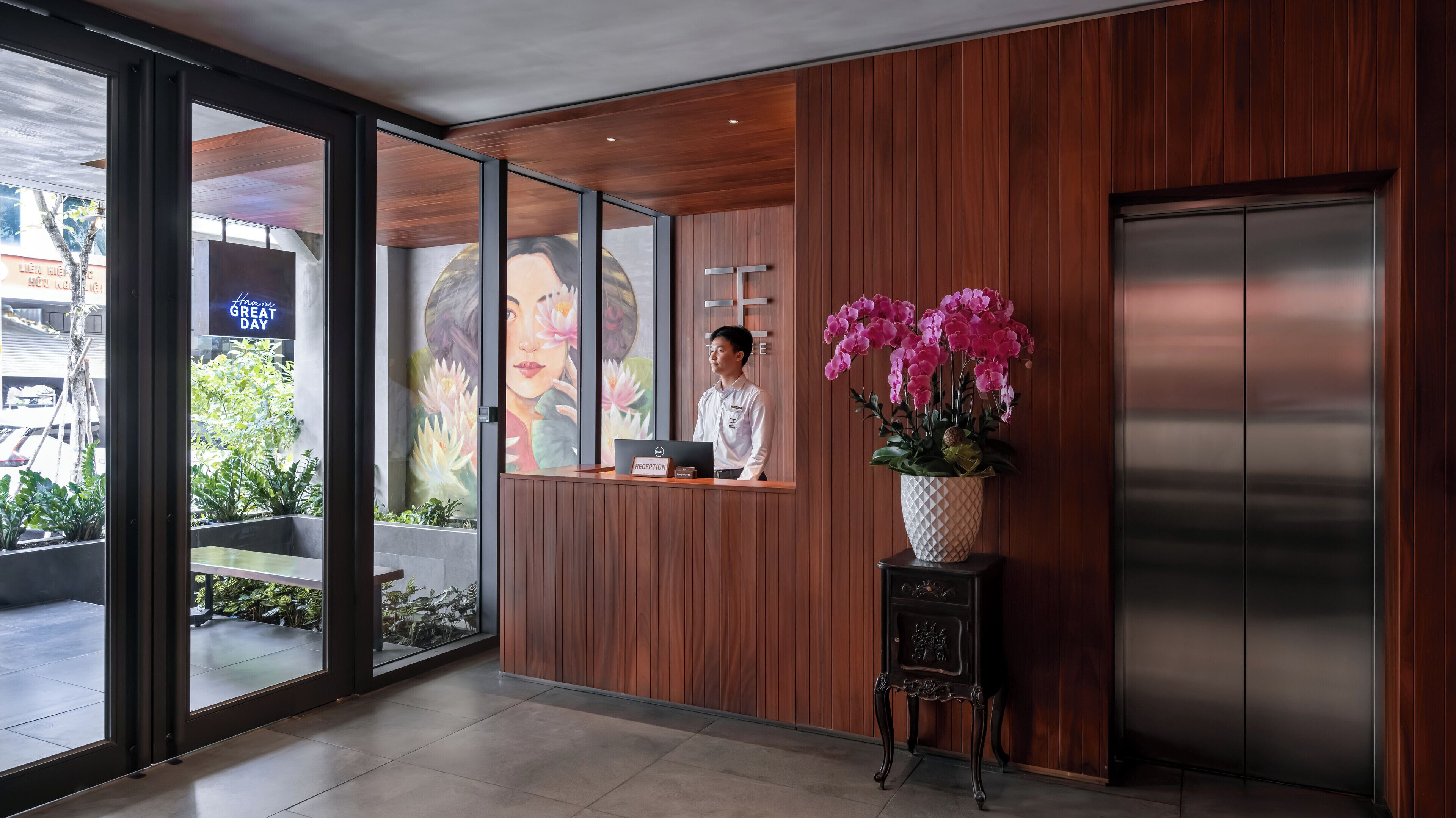 Un mostrador de recepción con una computadora portátil, un florero con flores rosas y un ventanal con vistas al exterior.