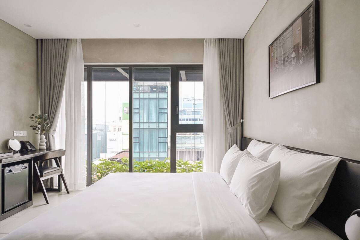 A modern hotel room with a large bed, a desk with a telephone, and a view of the city through large windows.