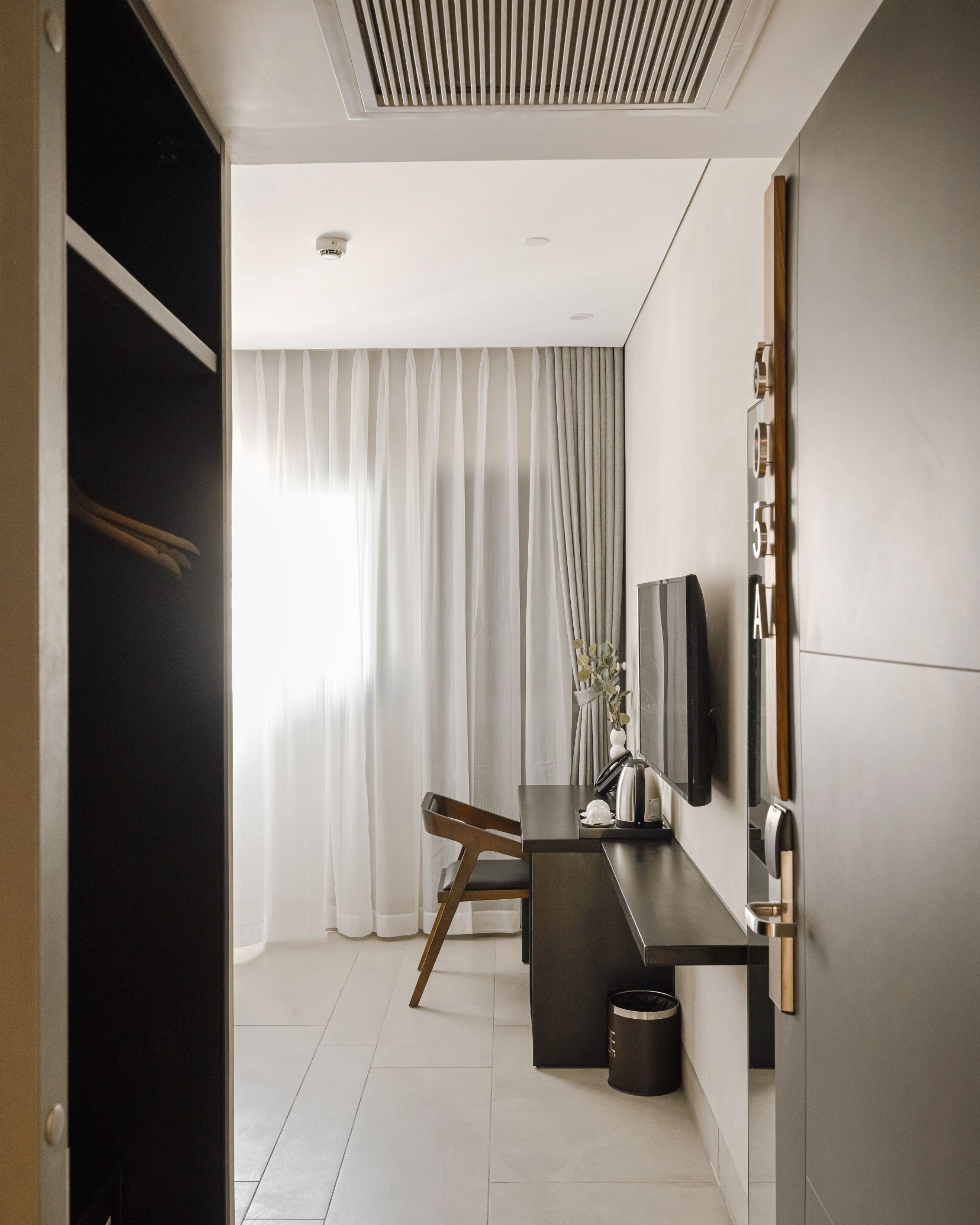 A modern hotel room with a large window, a desk with a chair, a flat-screen TV mounted on the wall, and a wardrobe.