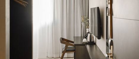 A modern hotel room with a large window, a desk with a chair, a flat-screen TV mounted on the wall, and a wardrobe.