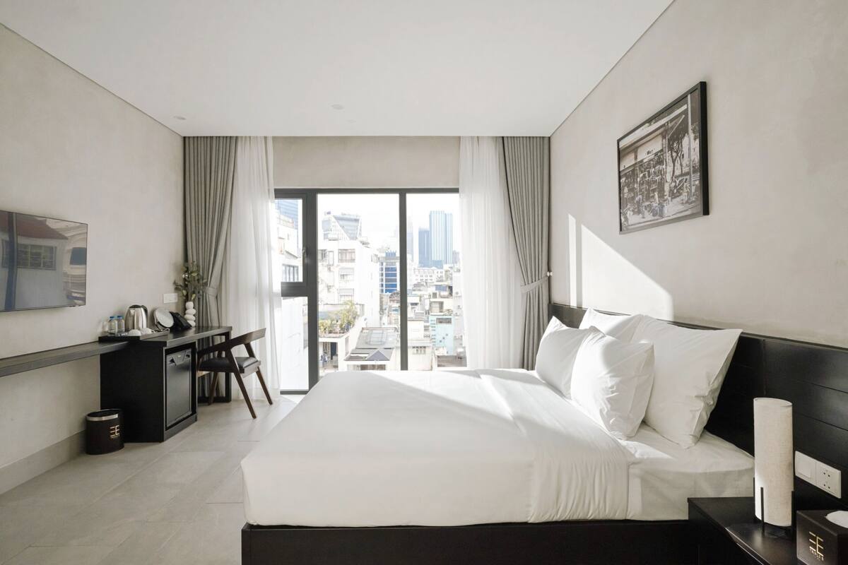 A modern hotel room with a large bed, a desk with a chair, a small kitchenette, and a city view through the window.