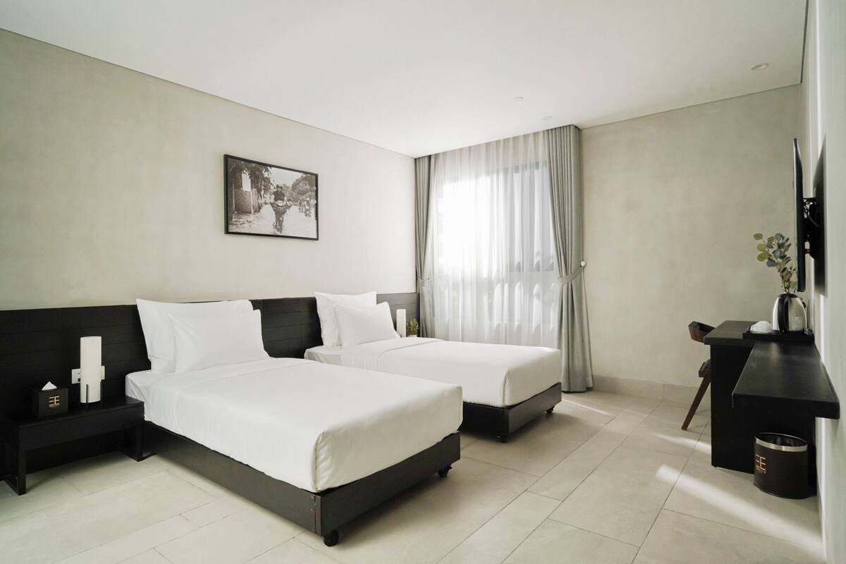 A hotel room with two beds, a desk, a chair, a TV, and a window with curtains.