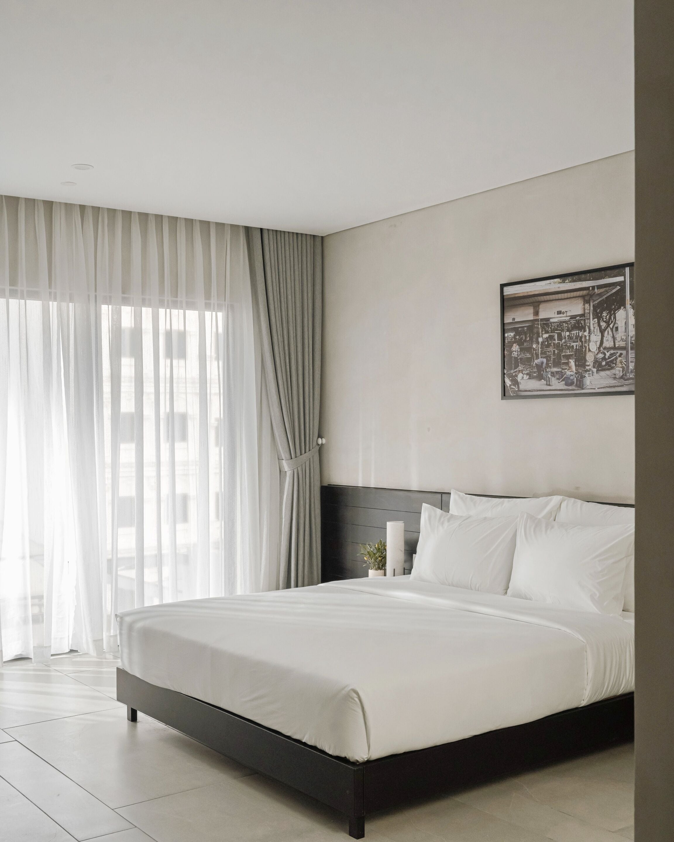 A modern hotel room with a large bed, a headboard, a framed picture on the wall, and sheer curtains on the windows.