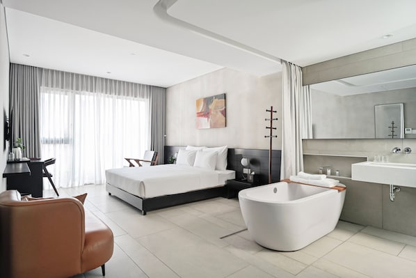 A modern hotel room with a large bed, a sitting area, a bathtub, and a bathroom with a sink and mirror.