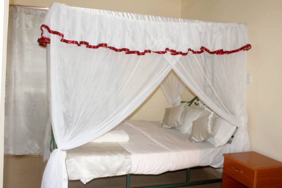 A single bed with a white canopy and red trim, a wooden nightstand, and a plain wall.