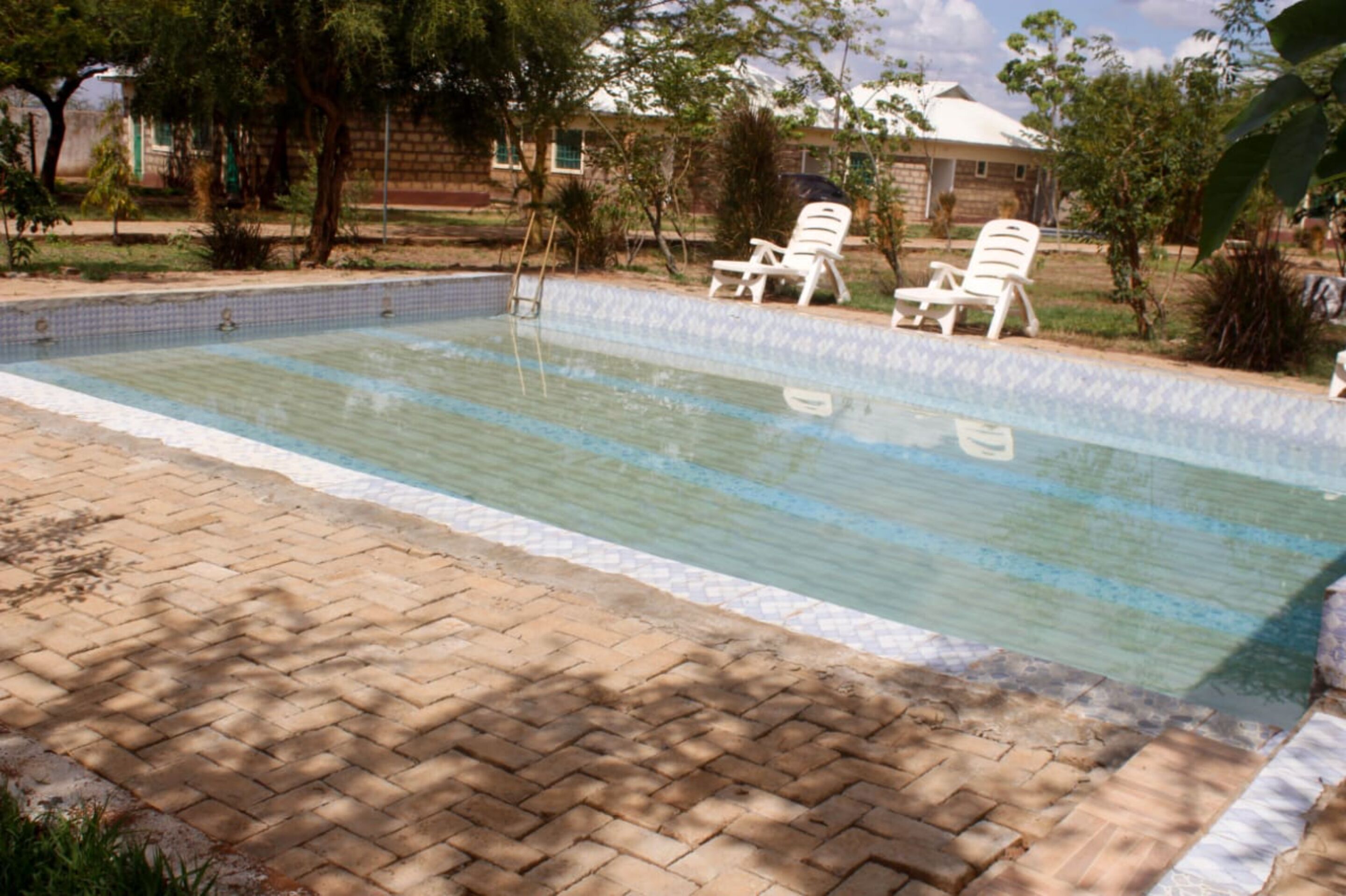 Una piscina con borde de azulejos, rodeada por un patio de ladrillo y dos sillones blancos.