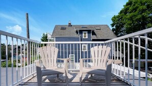 Eine Holzterrasse mit zwei Adirondack-Stühlen, die auf ein Haus mit dunklem Dach und Schornstein ausgerichtet ist.