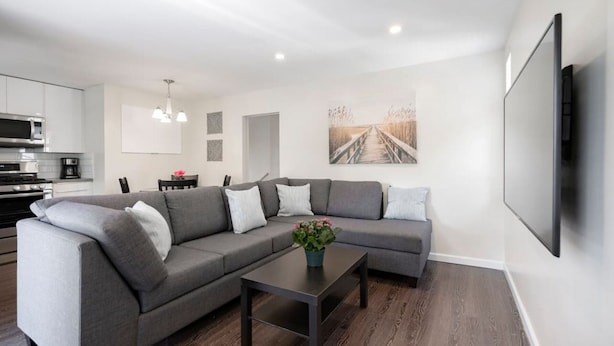 Ein modernes Wohnzimmer mit einem grauen Ecksofa, einem Couchtisch mit Pflanze, einem Essbereich mit Stühlen und einer Küche im Hintergrund.