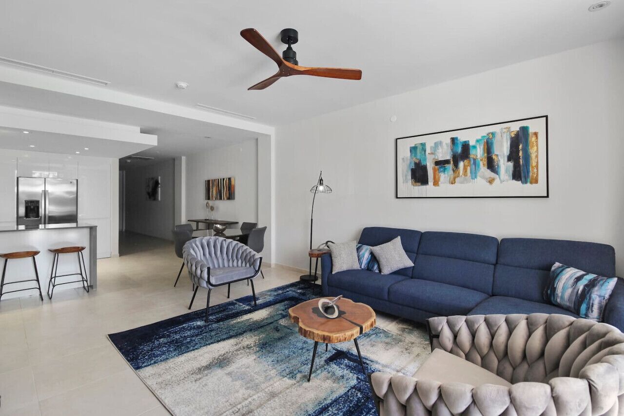 A modern living room with a blue sofa, a wooden coffee table, and a tufted armchair. A ceiling fan is visible above.