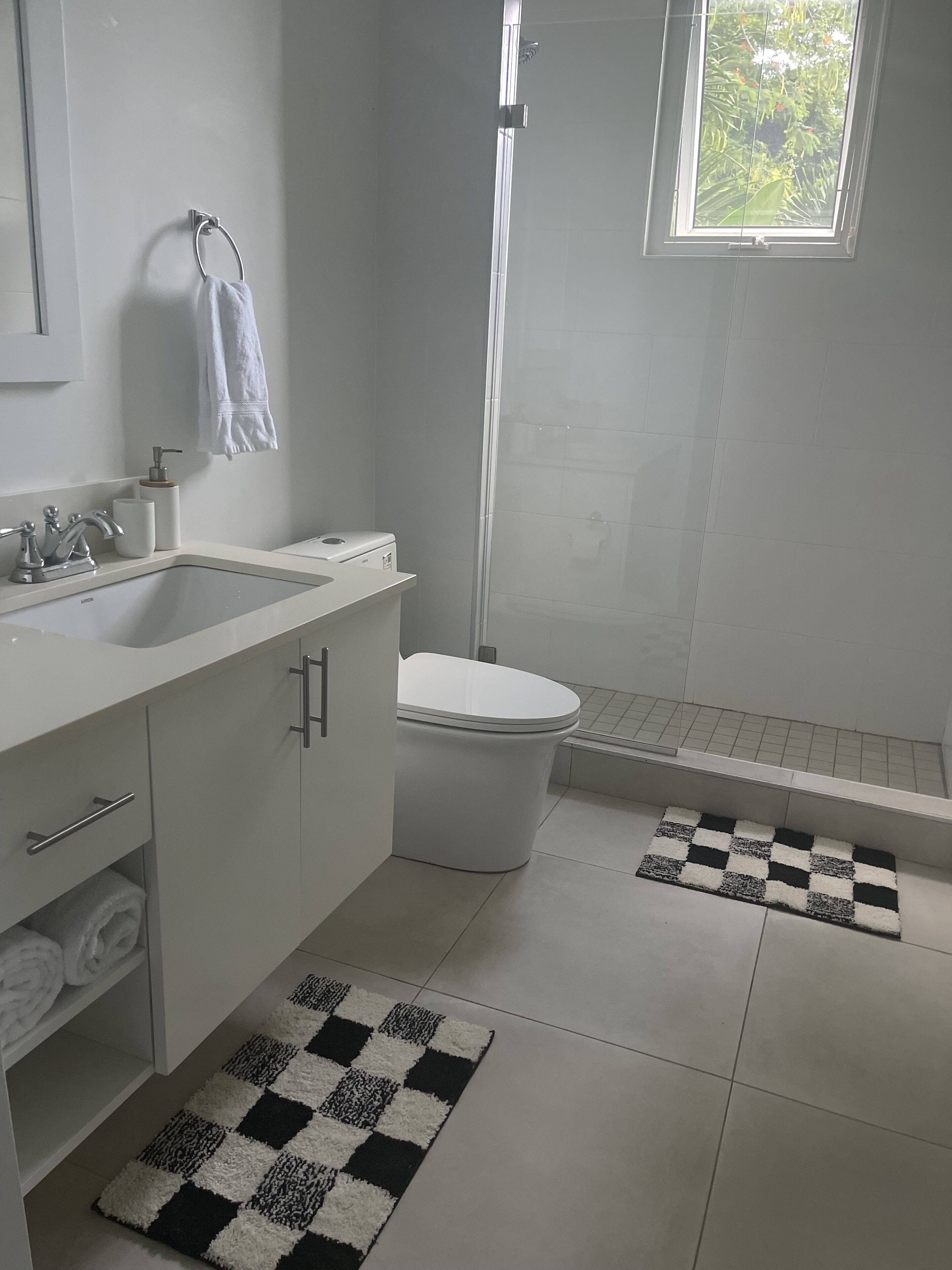 A modern bathroom with a white sink, toilet, and shower. There are two black and white checkered rugs on the floor.