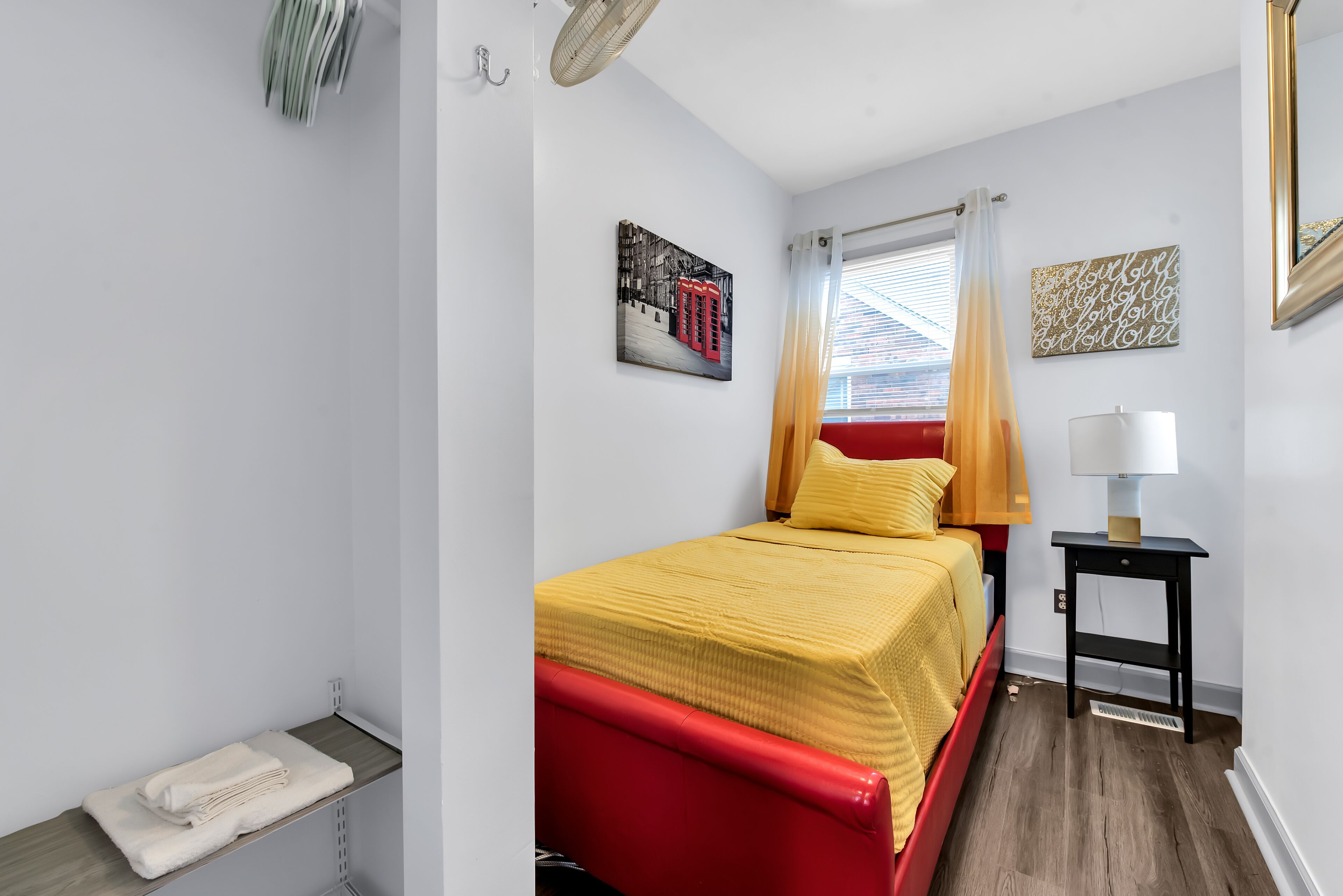 Ein Schlafzimmer mit einem roten Bett, gelber Bettwäsche, einem Nachttisch mit Lampe und einem Fenster mit Vorhängen.
