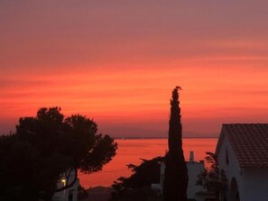 Ein Sonnenuntergang über ruhigem Gewässer, Bäume und ein Gebäude mit Ziegeldach.