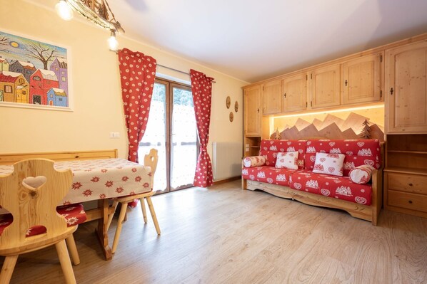 A cozy room with a wooden table and chair, a red sofa with heart patterns, wooden cabinets, and a painting of houses on the wall. - Nice apartment in the immediate vicinity of cable car (Canazei)