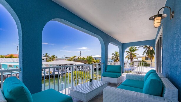 A balcony with white wicker furniture, a pool, and a view of palm trees and buildings. - Sandal Escape: 6BR Canal Front Oasis w/ Heated Pool, Dog-Friendly, Dockage & Fast Atlantic Access (Marathon)