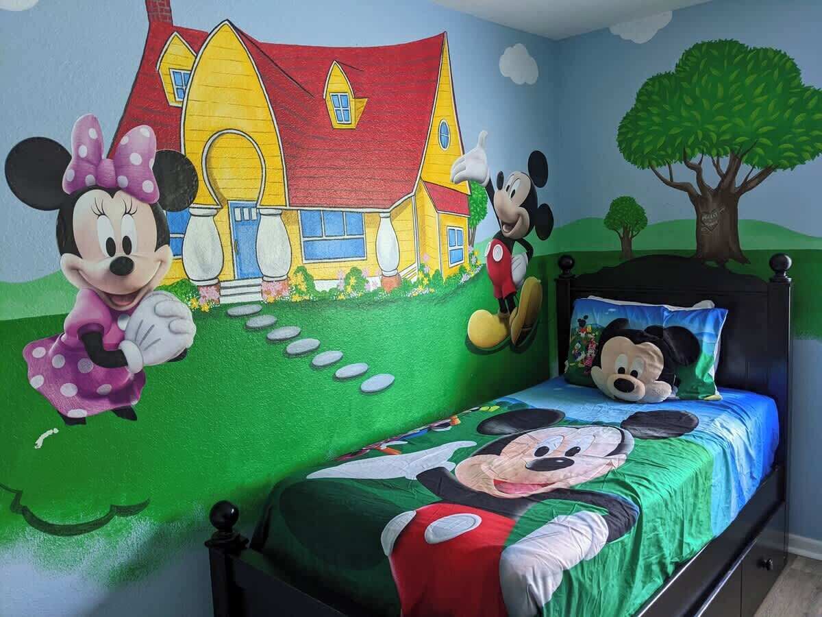 Una habitación infantil con decoración de Mickey Mouse y Minnie Mouse, una cama con almohada de Mickey Mouse, y un mural de una casa y un árbol.