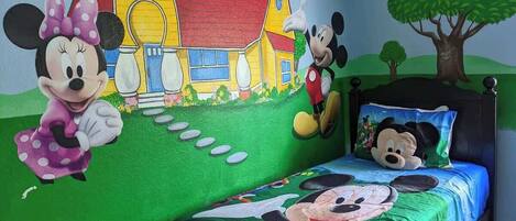 Una habitación infantil con decoración de Mickey Mouse y Minnie Mouse, una cama con almohada de Mickey Mouse, y un mural de una casa y un árbol.