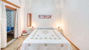 Un dormitorio con cama, dos mesitas de noche, un tocador, un espejo, una puerta con cortinas estampadas y una pared con un cuadro enmarcado.