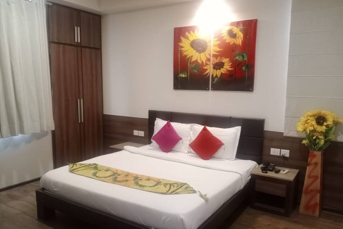 A hotel room with a bed, a nightstand, a vase with sunflowers, and a large painting of sunflowers on the wall.