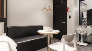 Una habitación de hotel moderna con un sofá negro, una mesita redonda pequeña con una caja de pañuelos, una silla blanca, un ventilador de pie y una puerta con un picaporte rojo.