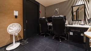 Una habitación pequeña y moderna con un muro a rayas negras y blancas, un escritorio con una computadora, dos sillas de oficina negras, un refrigerador pequeño y un ventilador de pie.