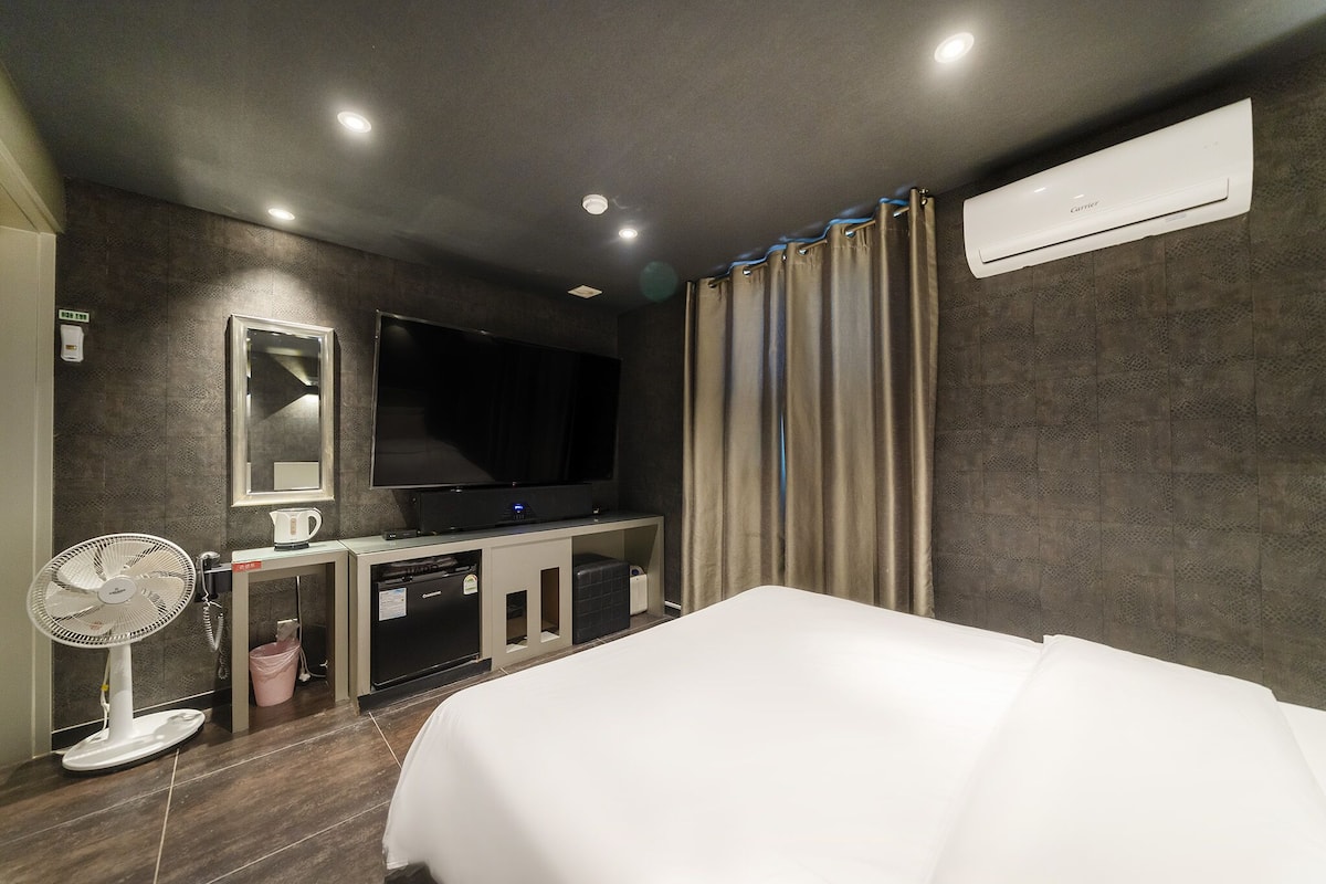 A hotel room with a bed, a flat-screen TV, a fan, a coffee maker, and an air conditioning unit.