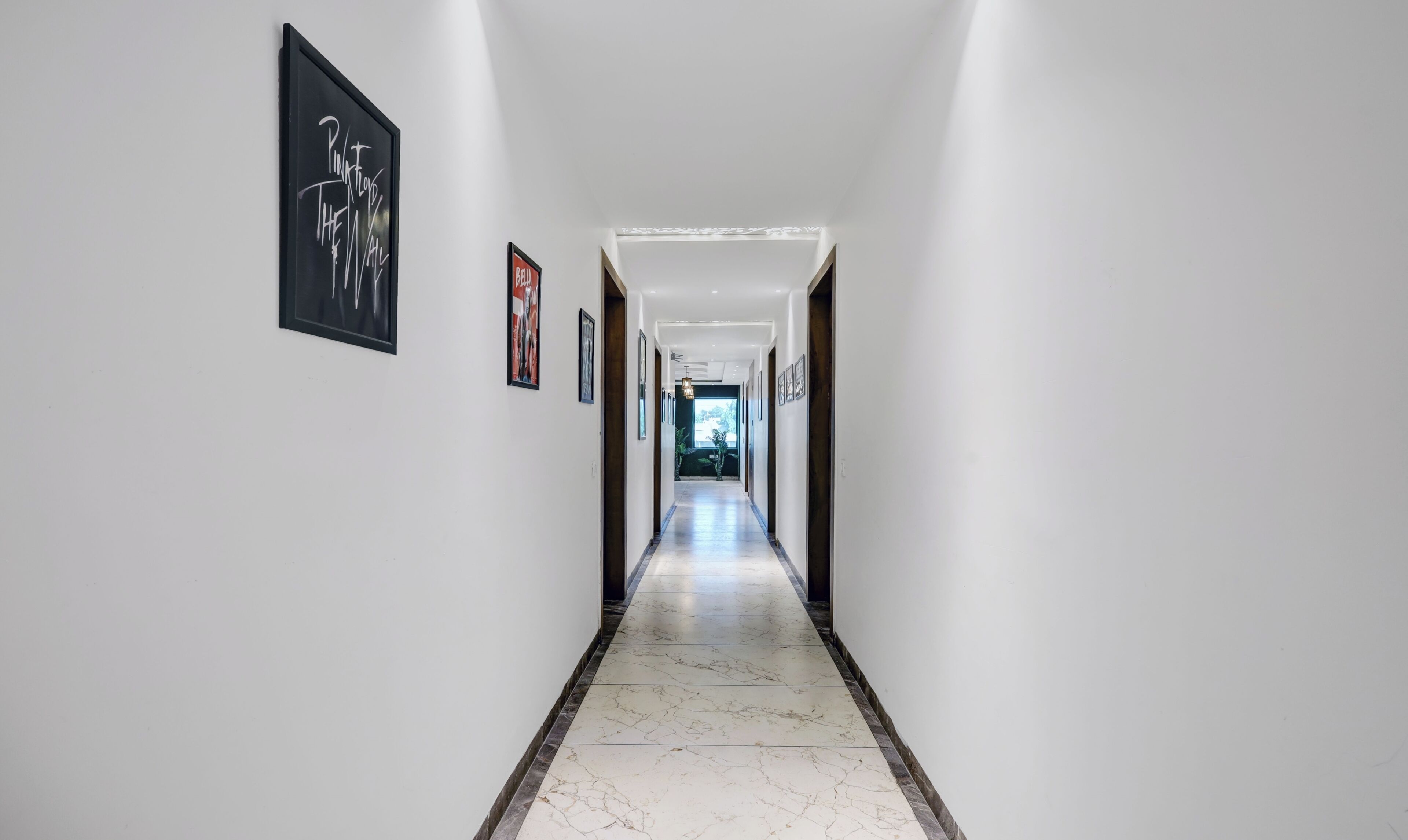 A long, narrow hallway with white walls and a marble floor. There are framed pictures on the walls and a door at the end of the hallway.