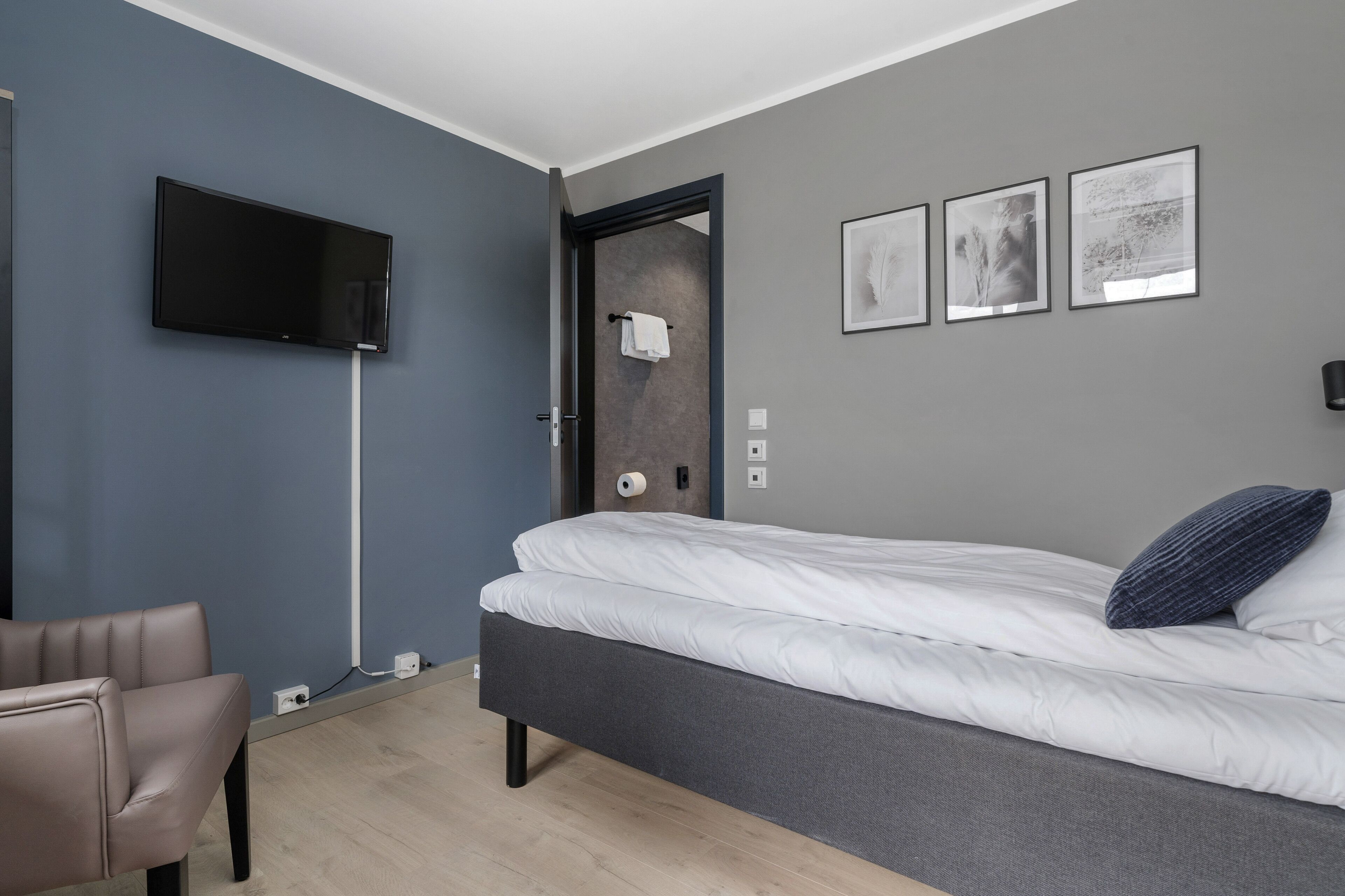 Quarto com cama, televisão de tela plana na parede, uma cadeira e porta que dá para um banheiro.