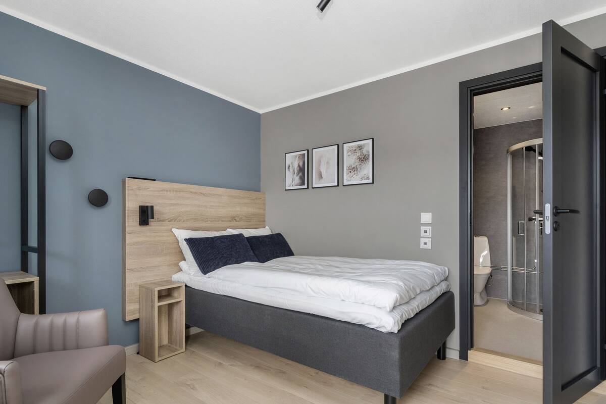 Un dormitorio moderno con cama gris, cabecero de madera y una mesita de noche pequeña. Hay tres cuadros enmarcados en la pared y una puerta que da a un baño.