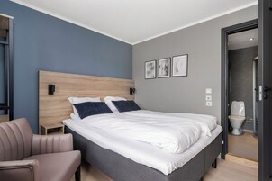 Un dormitorio moderno con una cama grande, una mesita de noche, una silla y un baño visible a través de una puerta abierta.