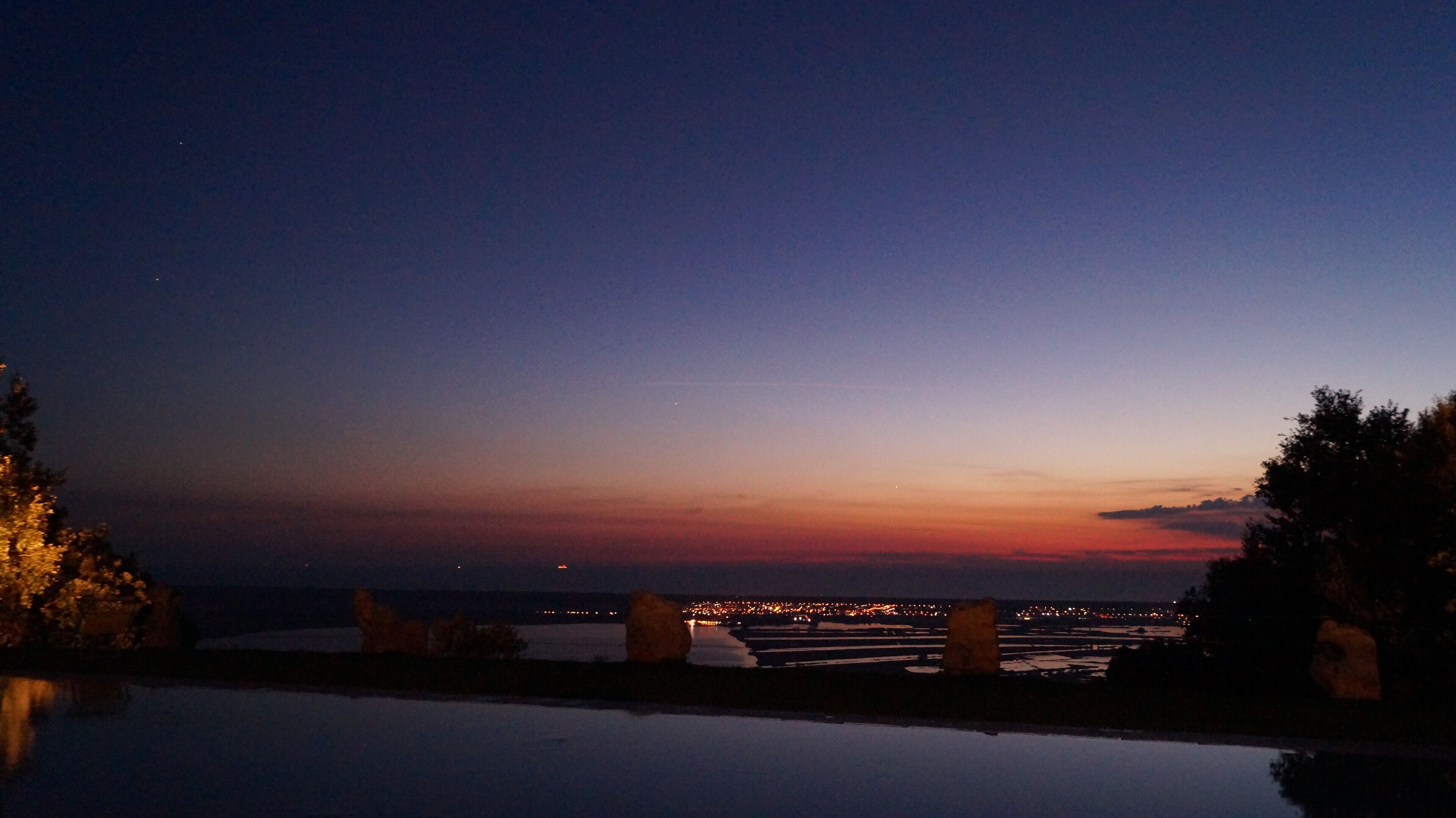 Ein Nachtblick auf eine Küstenregion mit ruhigem Gewässer, das Lichter vom Ufer reflektiert.