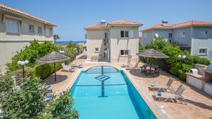 A swimming pool with lounge chairs and umbrellas, surrounded by buildings with a Mediterranean architectural style.